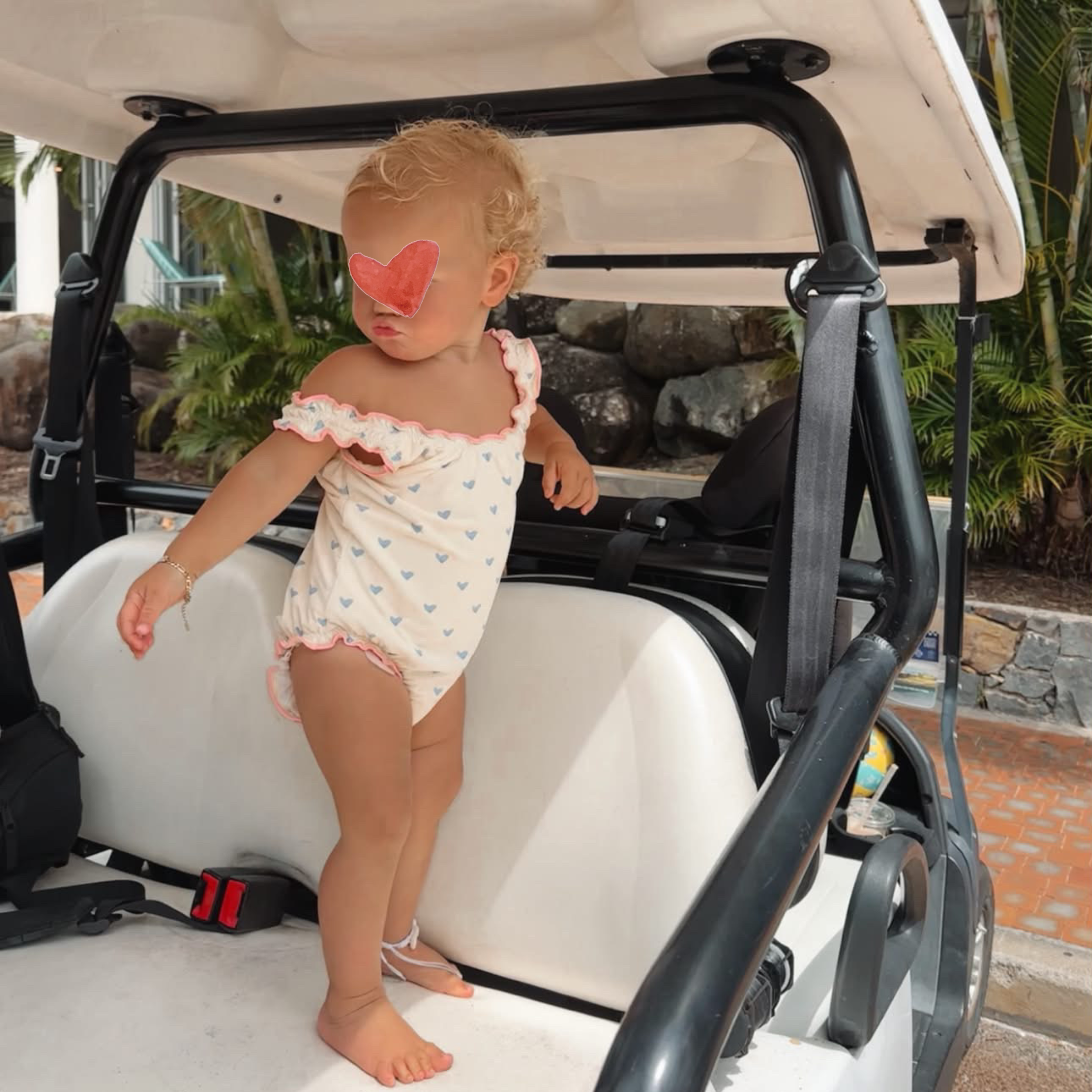 A toddler stands barefoot in the back of a white golf cart, wearing a white off-the-shoulder one-piece swimsuit with small blue hearts and pink ruffle trim. The child has a pink heart-shaped sticker over their eyes, with palm trees and rocks in the background.