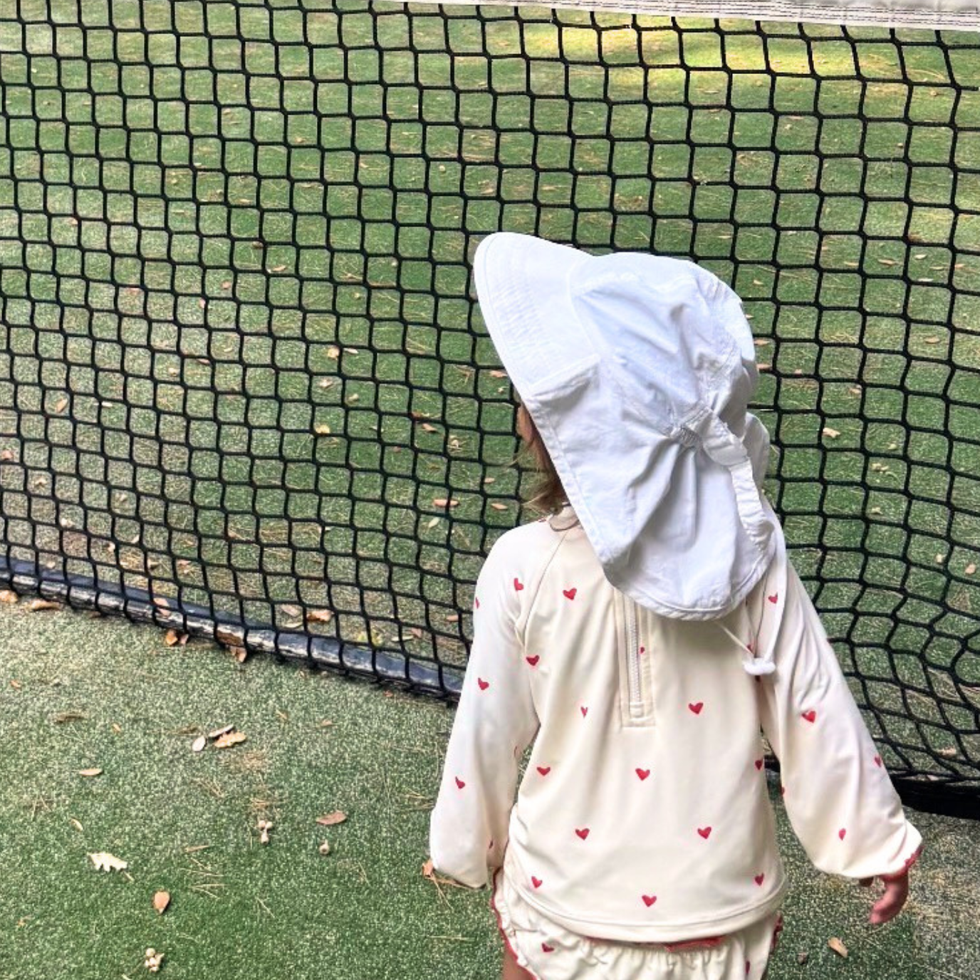 A toddler stands on an outdoor tennis court behind a net, wearing a cream long-sleeve romper with small red heart prints and a matching sun hat with a tie-back.