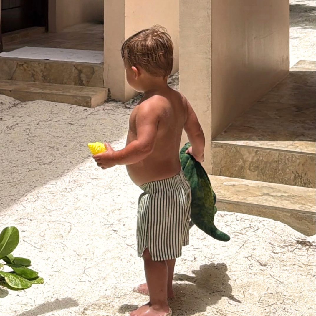 A toddler standing outdoors on sand in front of beige stucco columns, wearing green and white vertically striped swim trunks and holding a yellow cup and a green dinosaur toy.