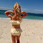 A young girl with blonde hair in a ponytail stands on a sandy beach facing the ocean, wearing a pale yellow ruffled two-piece swimsuit.