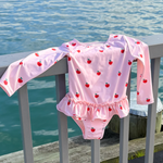 A pink long-sleeve baby swimsuit with a red apple print and ruffled hem, draped over a gray wooden dock railing with water in the background.