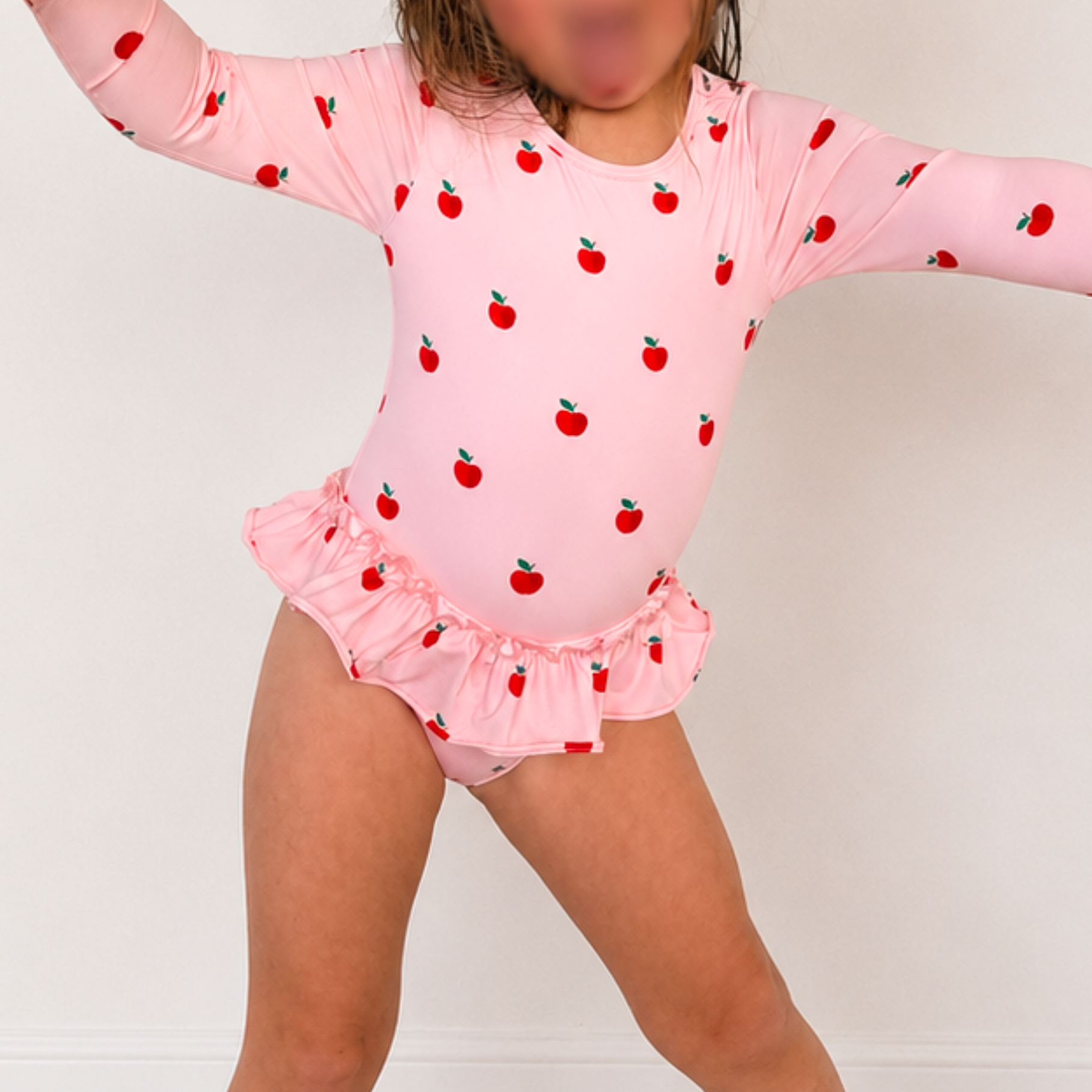 A young child wearing a pink long-sleeve leotard with a red apple print and a ruffled bottom, posed against a plain light background.