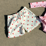 White swim trunks with a red apple print pattern, laid flat on a gray concrete surface next to a pink swimsuit.