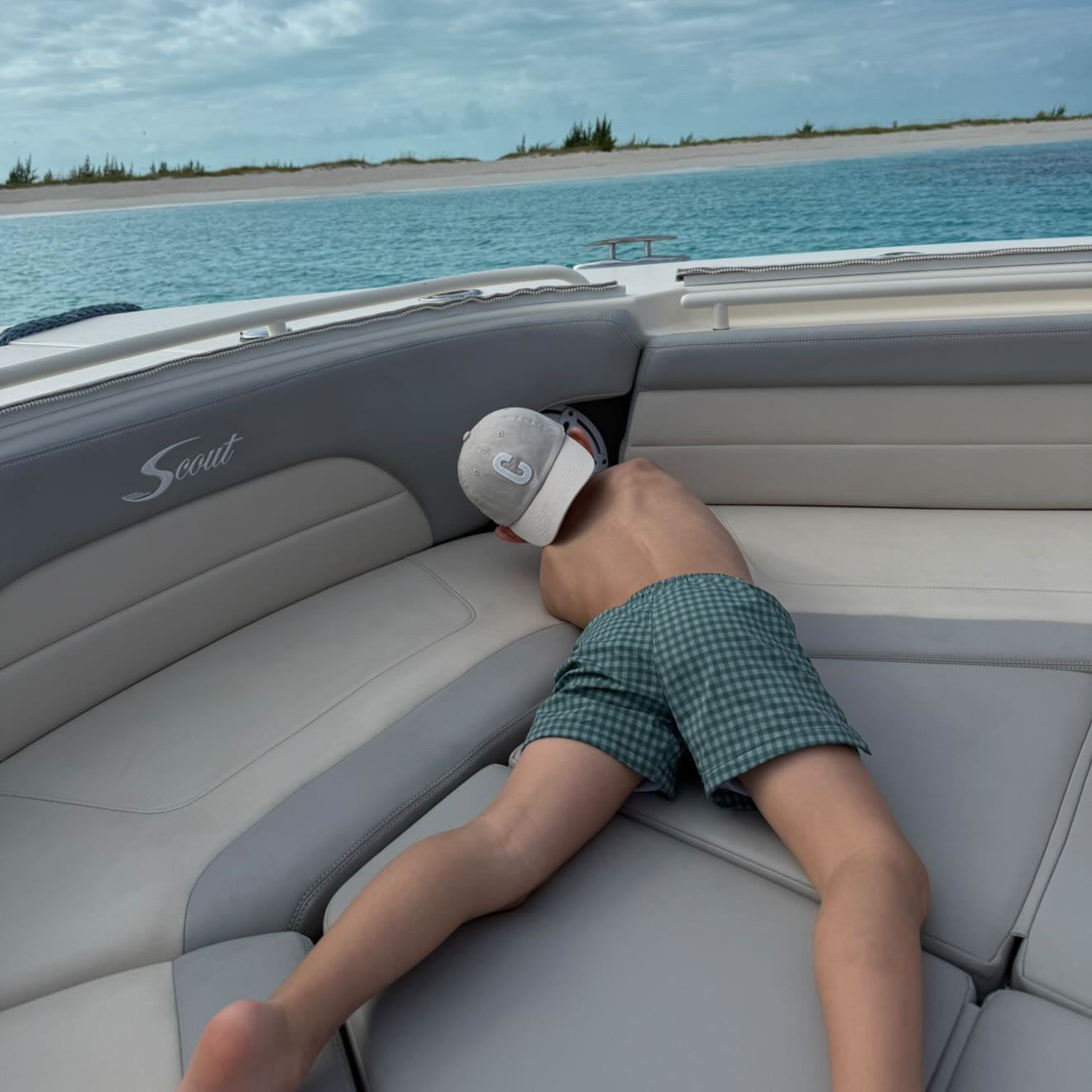 A person lying on the cushioned seating of a boat, wearing green and white checkered swim trunks and a white cap, with blue water and sky in the background.