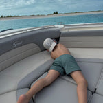 A person lying on the cushioned seating of a boat, wearing green and white checkered swim trunks and a white cap, with blue water and sky in the background.
