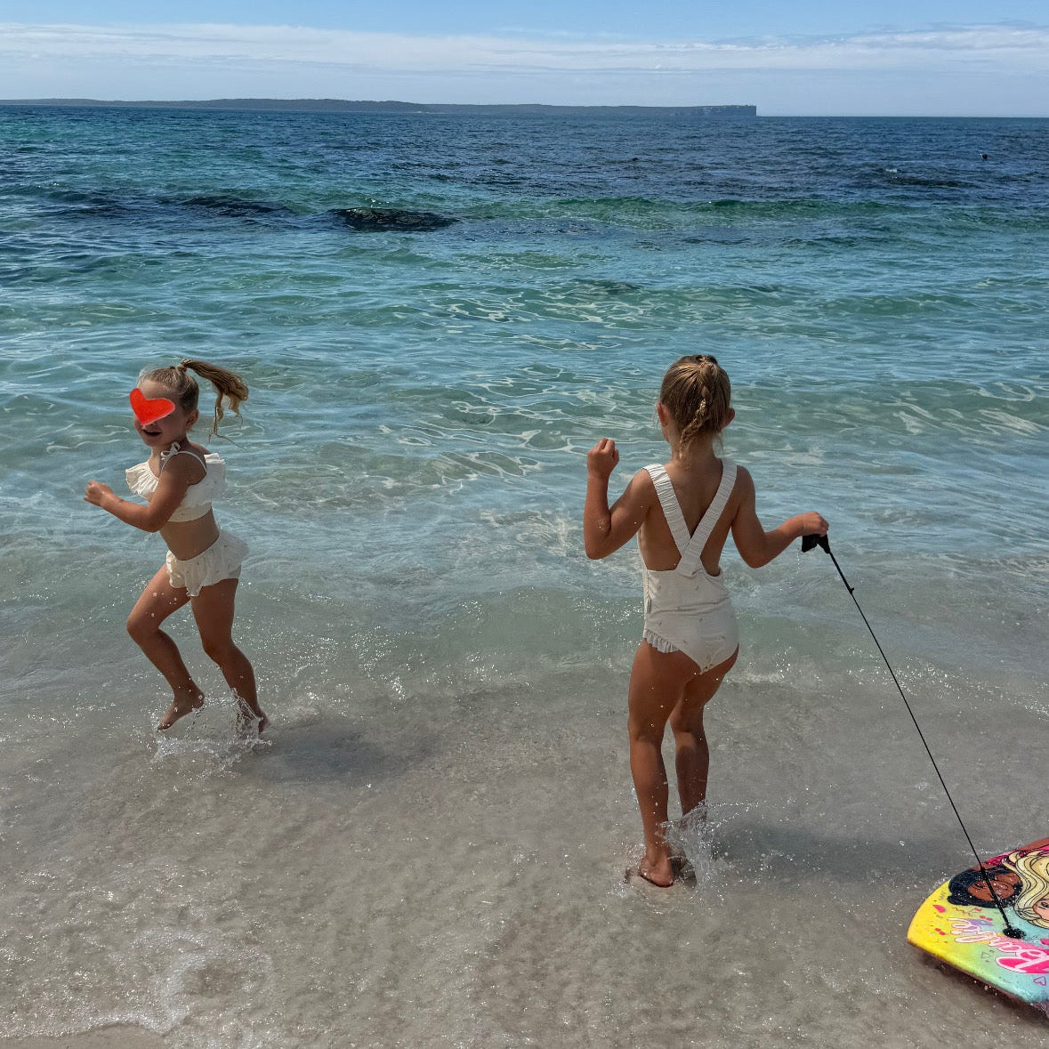 Two girls in white swimwear running in shallow ocean water on a sandy beach, with one holding a leash attached to a colorful board. The background features a bright blue sky with white clouds and turquoise water.