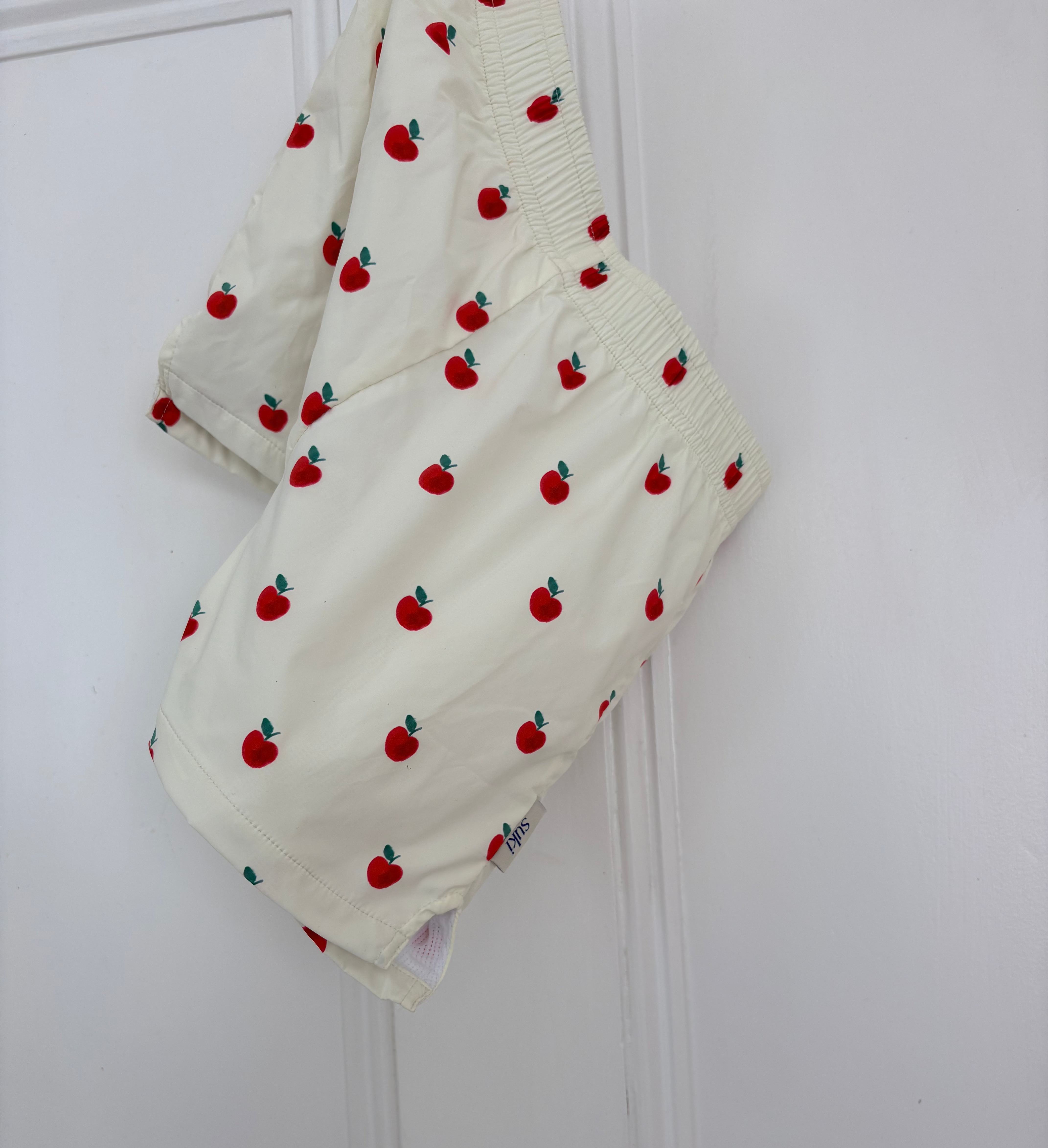 Off-white baby shorts with a red apple print, hanging on a white door.