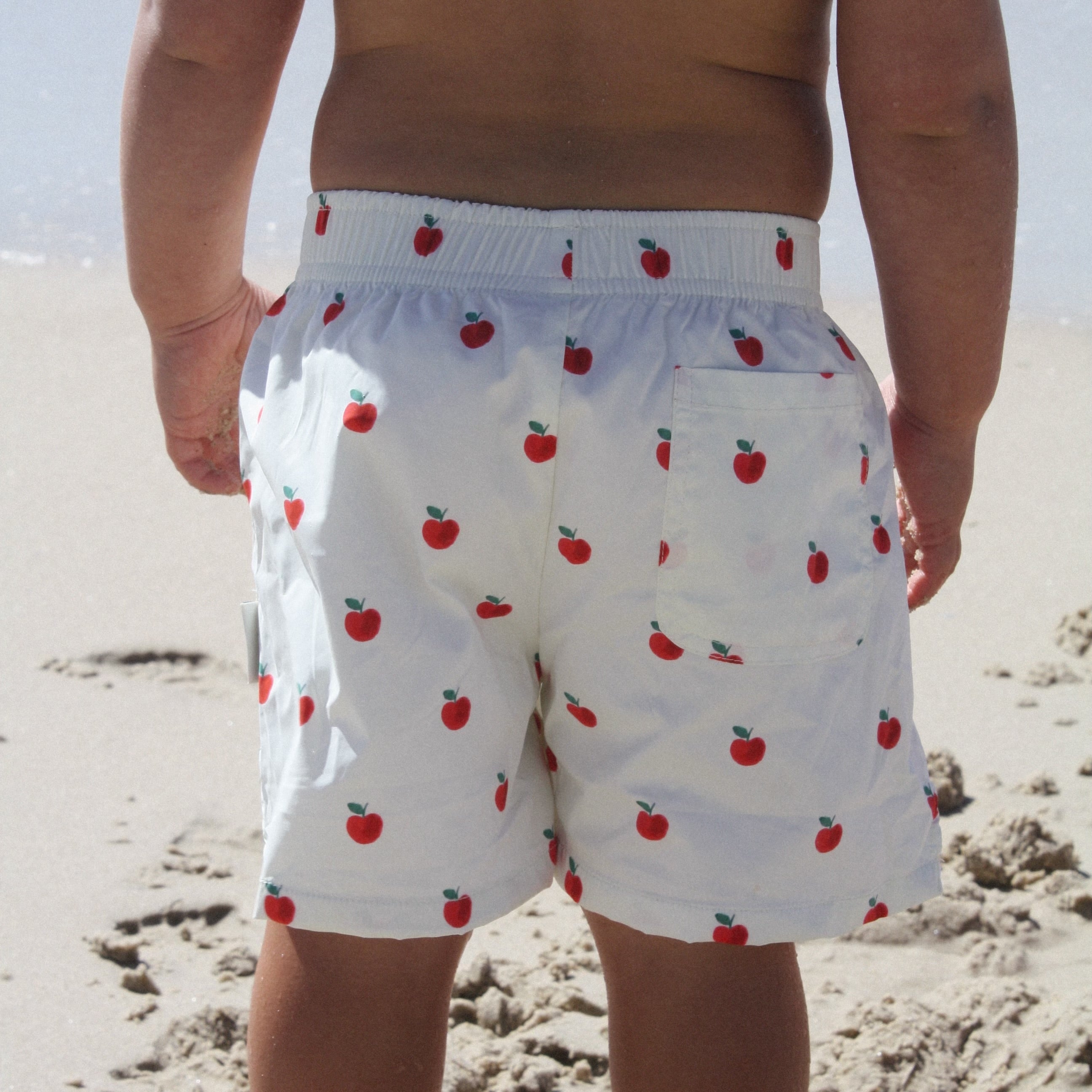 A child viewed from behind on a sandy beach, wearing white swim trunks with a red apple print.
