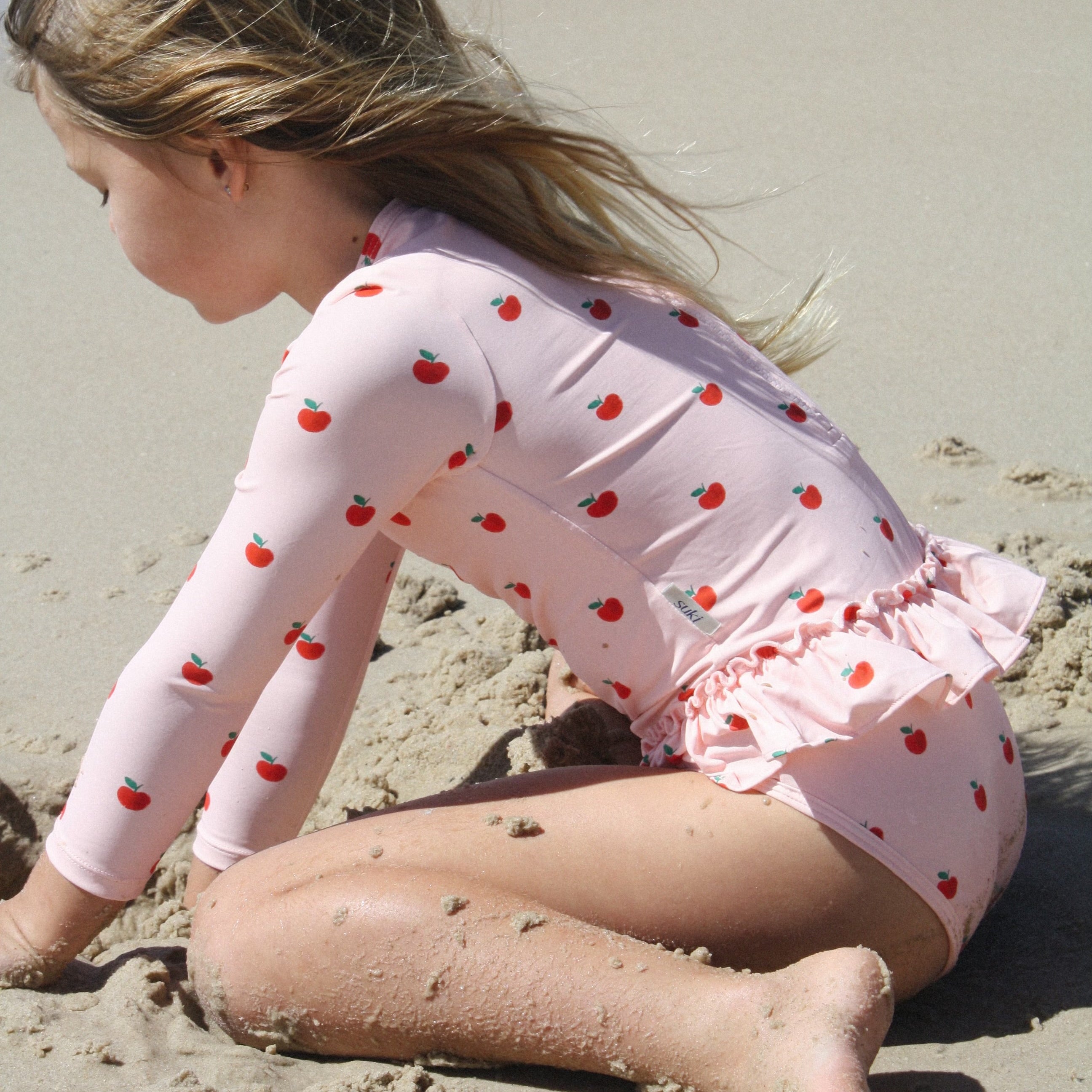 A young girl with blonde hair sitting on a sandy beach, wearing a light pink long-sleeve swimsuit with a red strawberry pattern.