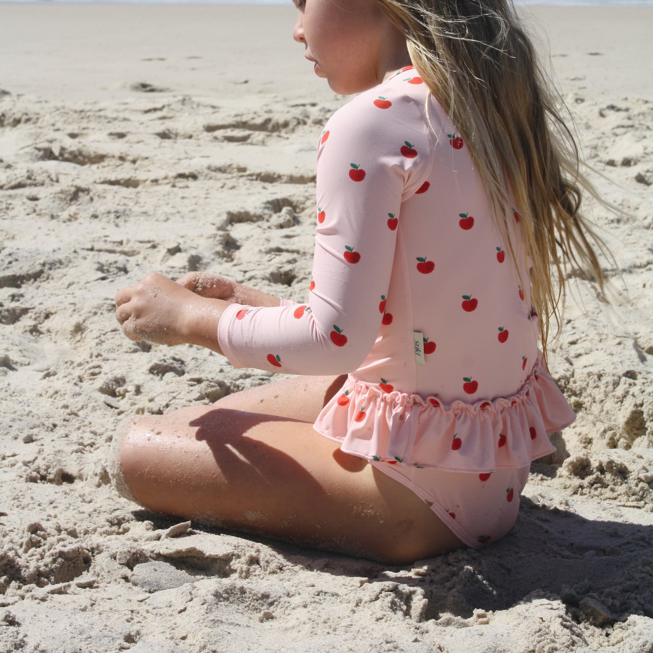A young girl with long blonde hair sits on a sandy beach, wearing a light pink long-sleeve swim shirt and matching ruffled swim bottoms with a red cherry print. The ocean waves are visible in the background.