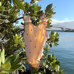 A cream-colored baby romper with small red hearts and red ruffle trim hanging from a green tree branch outdoors, with water and a bridge in the background.