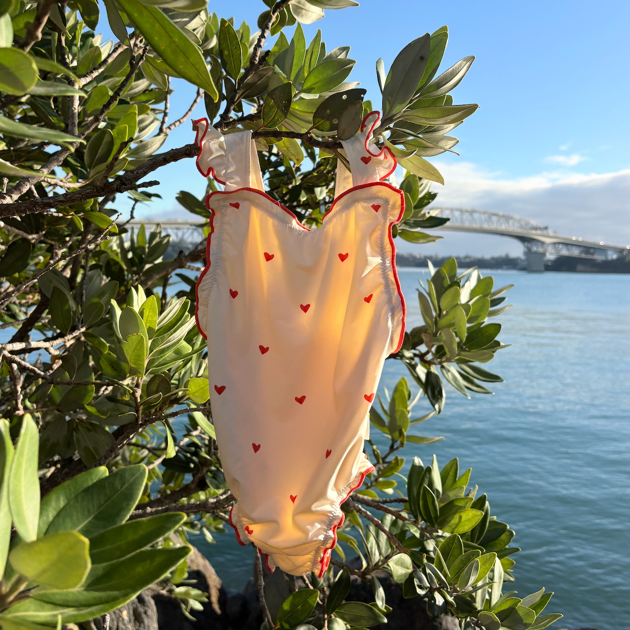 A cream-colored baby romper with small red hearts and red ruffle trim hanging from a green tree branch outdoors, with water and a bridge in the background.