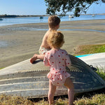Two children in swimwear standing on and beside a overturned metal boat on grass by a sandy shoreline, with water, distant boats, and a tree canopy in the background.