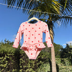 A pink long-sleeve baby swimsuit with a red cherry print, hanging on a wooden hanger outdoors against a background of palm trees and blue sky.