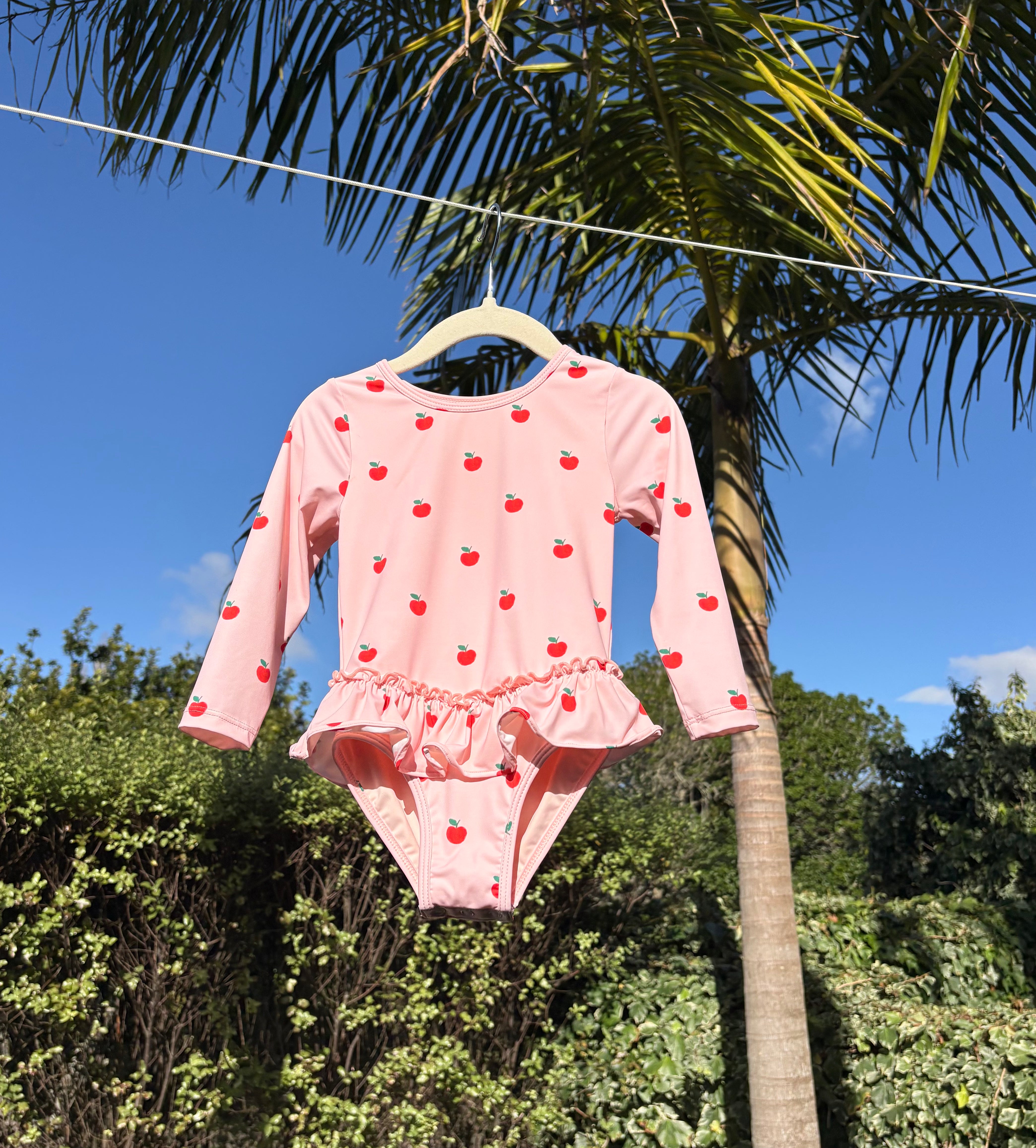 A pink long-sleeve baby swimsuit with a red cherry print, hanging on a wooden hanger outdoors against a background of palm trees and blue sky.