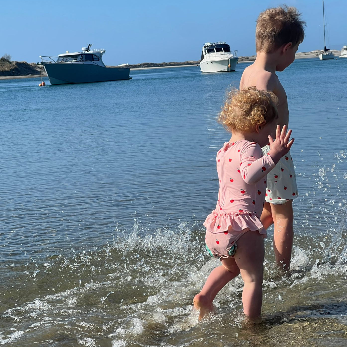 A young child in a pink polka-dot swimsuit walks in shallow water while an older child stands beside them in a sunny outdoor harbor scene with boats in the background.