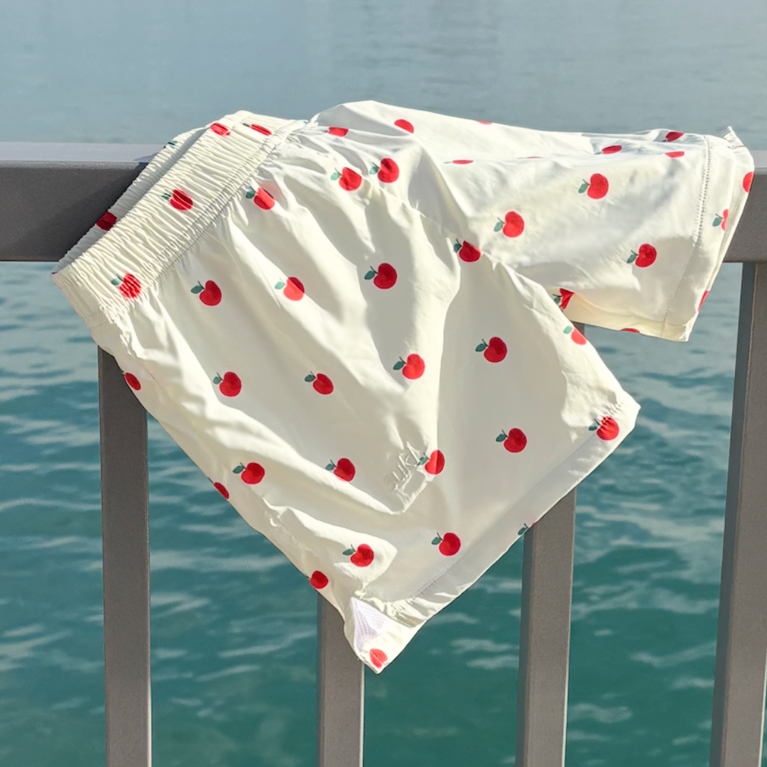 White swim trunks with a red apple print, draped over a gray metal railing with a body of water in the background.